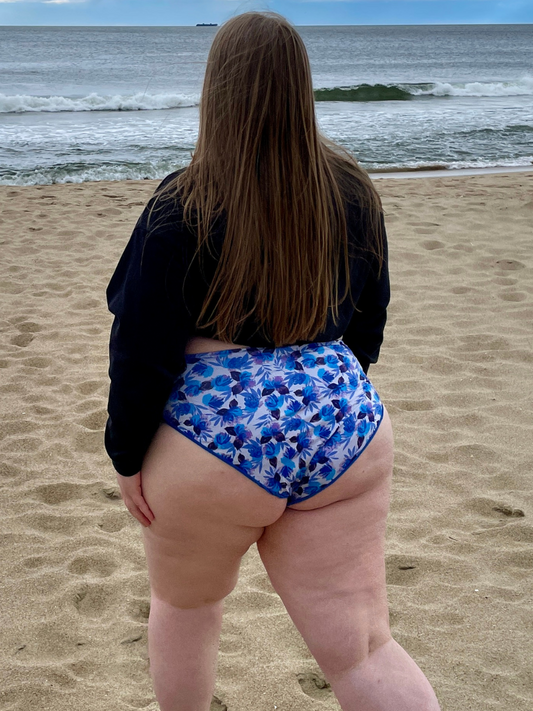 woman on the beach in a cropped black crewneck sweatshirt and high waisted, high hip, blue floral bikini bottoms