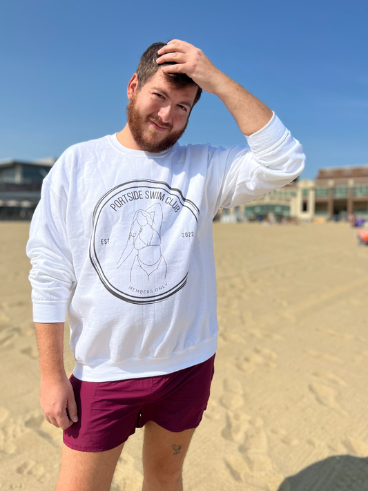 man on the beach in white crewneck with a circular graphic that has a line drawing of a girl in a bikini and says "Portside Swim Club - Members Only"