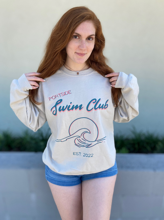 woman in a long sleeve, sand colored crewneck with a graphic of a wave in front of a circle with the words "Portside swim club" above it