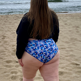 woman on the beach in a cropped black crewneck sweatshirt and high waisted, high hip, blue floral bikini bottoms