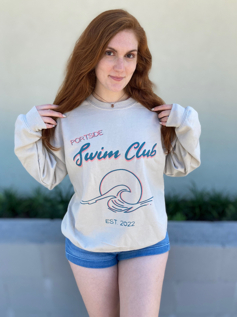 woman in a long sleeve, sand colored crewneck with a graphic of a wave in front of a circle with the words "Portside swim club" above it