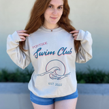woman in a long sleeve, sand colored crewneck with a graphic of a wave in front of a circle with the words "Portside swim club" above it