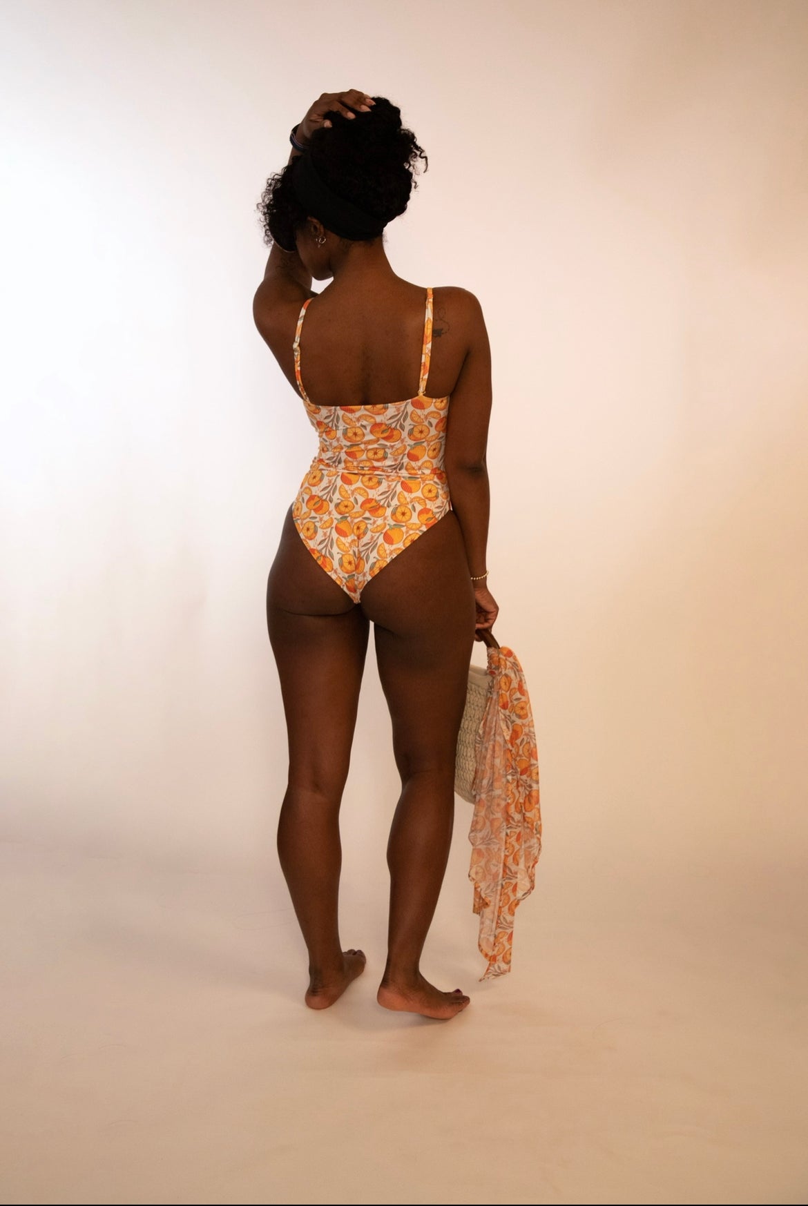 back of woman in reversible orange print one piece bathing suit holding a bag with a matching sarong tied around the handle